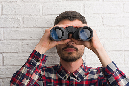 Young adult man over brick wall looking through binoculars with a confident expression