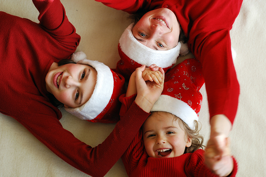 Picture of three children wearing red sweaters and santa hats laying down on the ground laughing with each other