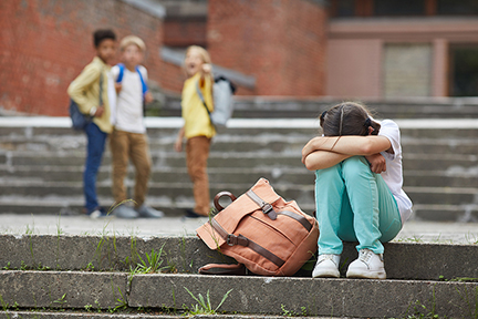crying schoolgirl sitting on stairs outdoors with group of teasing children bullying her in background
